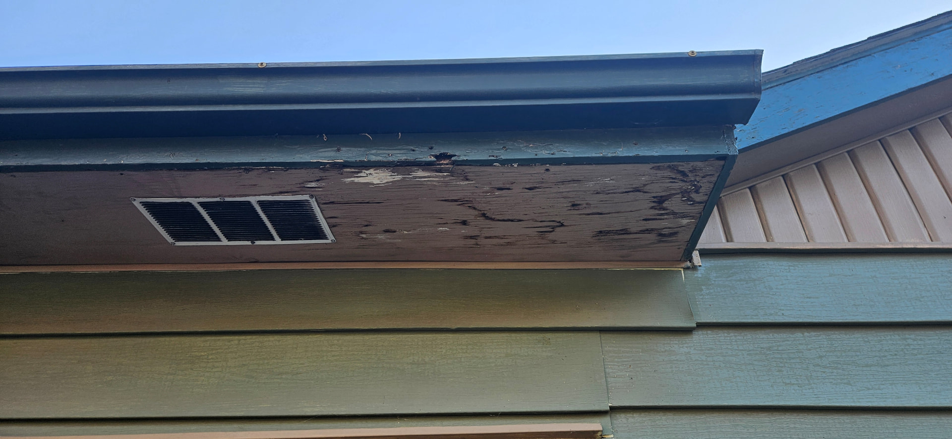 Close-up of storm wear on a Topeka home: dark gutter and fascia above a weathered wooden soffit with peeling paint and rot beside a louvered vent screen
