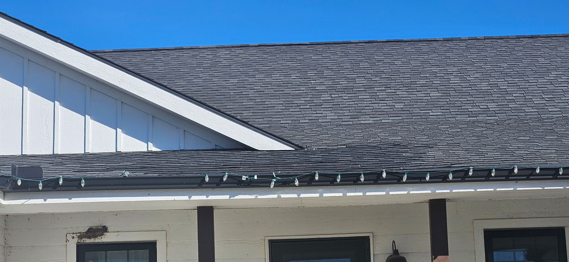 Roofline of a home with dark asphalt shingles above white board-and-batten siding, a black gutter lined with holiday lights, and a white porch below