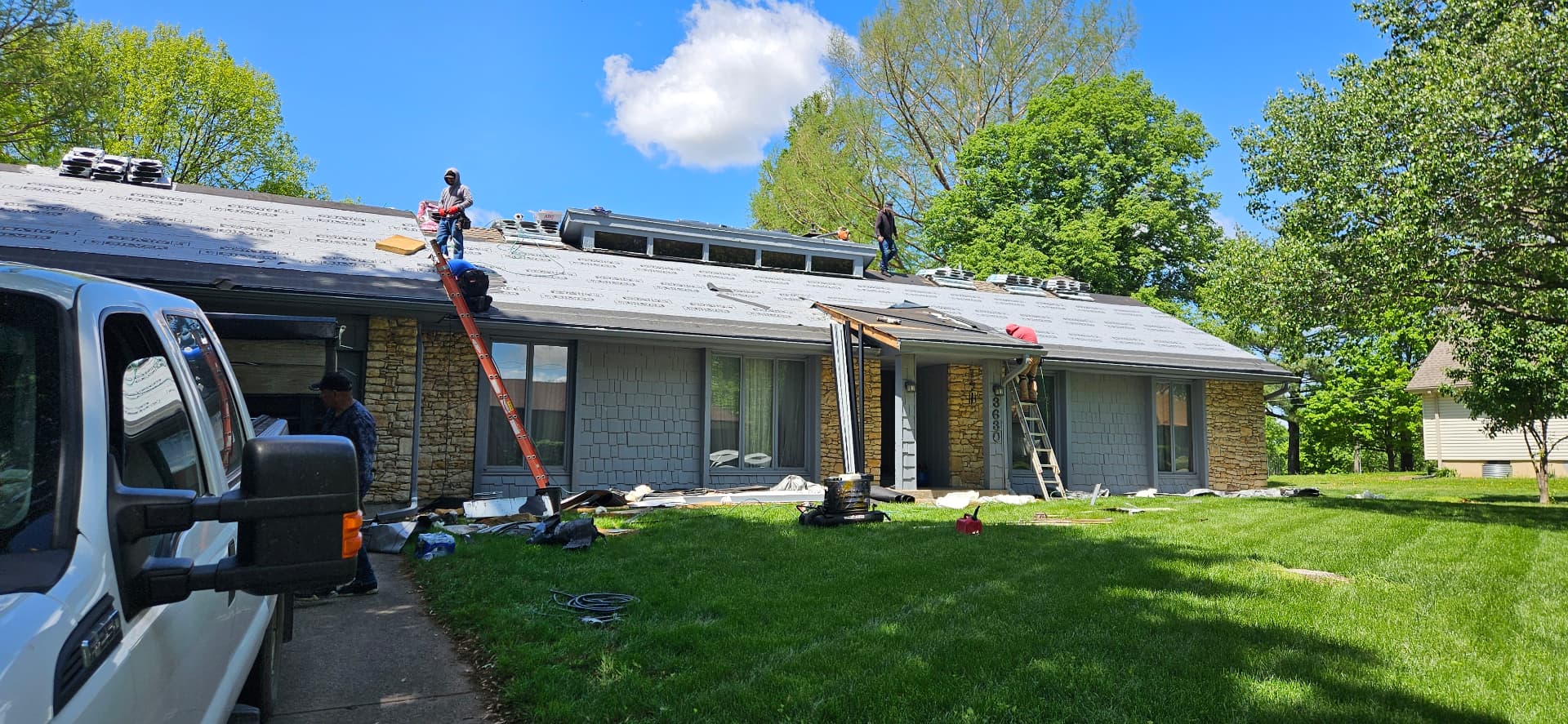 Kan Exteriors crew members replacing a roof on a Topeka home: grey synthetic underlayment laid over the deck, ladders at the eaves and a stone-and-shake exterior under a blue sky