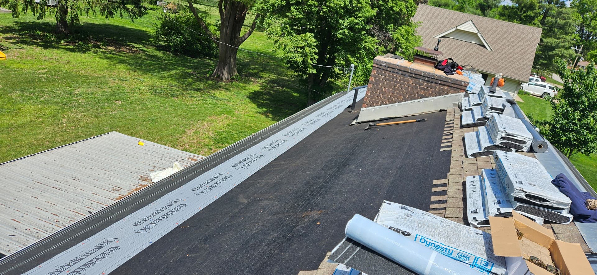 Kan Exteriors roof replacement in progress on a Topeka home: black underlayment over fresh decking, a brick chimney at the ridge and staged bundles of new asphalt shingles along the right side